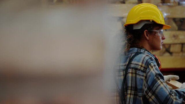 Framed through a blurred wooden post, a young female worker in a yellow hard hat and safety glasses sorts small wooden blocks inside a timber and pallet warehouse