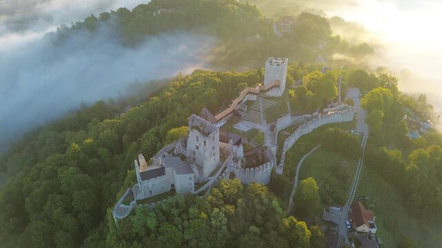 Aerial drone view of Celje Castle surrounded by lush forest, mist and warm golden sunlight, showcasing the medieval fortress complex on a wooded hill in Slovenia.
