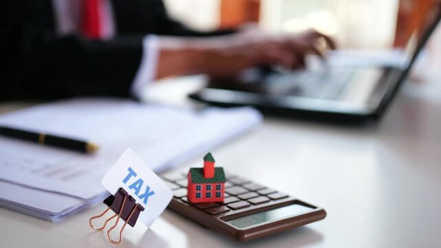 The word "Tax" and a miniature house are placed on a calculator on a desk, with an office worker typing on a laptop in the background. House and building tax. Property tax. Planning or budgeting for h