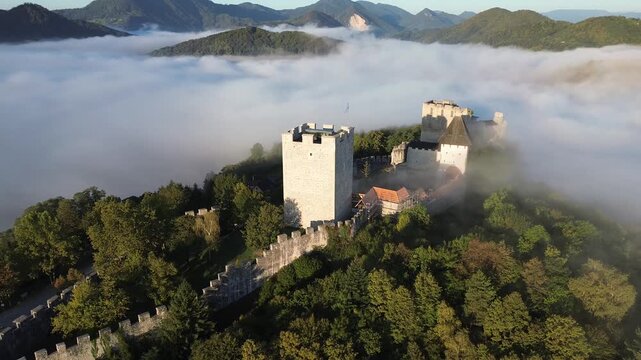 Aerial view of Celje Castle surrounded by fog in Slovenia, with medieval towers and walls emerging above the mist, autumn trees and mountain peaks in the background.