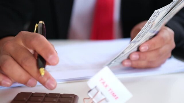 TAX TIME written on a clipped sheet of paper on a desk, representing the tax season concept, set against a background of an employee holding money and calculating figures. Concepts of tax payment plan