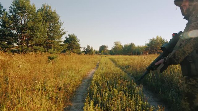 Soldier in ghillie suit walking on a dirt path through a grassy field with pine trees in background.