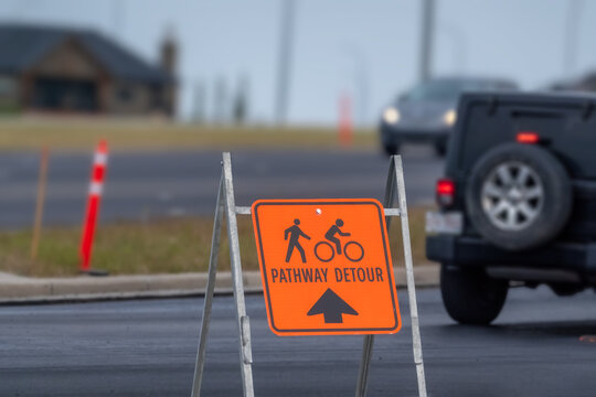 Orange pathway detour sign with pedestrian and cyclist symbols standing on a city street beside traffic.