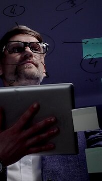 Mature male professional, wearing eyeglasses and suit, working late in a dark office, holding a digital tablet and brainstorming new business strategies with post it notes on a glass board