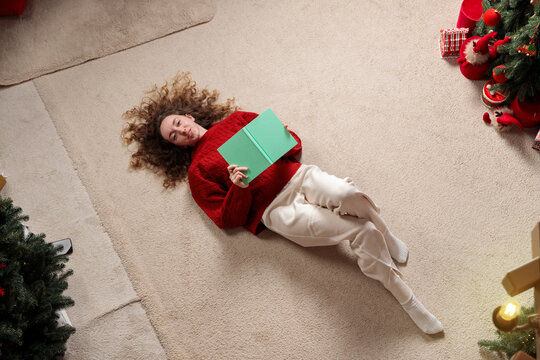 Woman wears a red sweater while lying on a carpeted floor surrounded by festive holiday decorations and evergreen trees