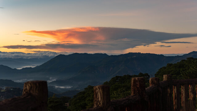 Dramatic sunset over layered mountain ridges with glowing orange clouds, misty valleys, and a rustic wooden railing framing the scenic highland landscape.
