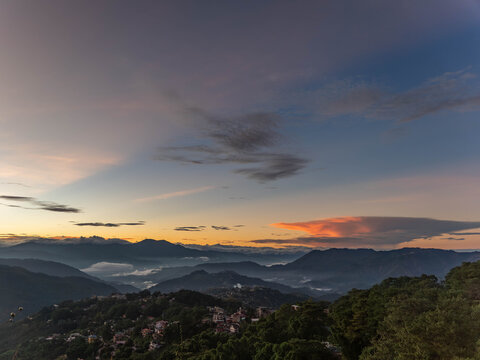 Panoramic mountain city landscape at sunset with layered ridges, misty valleys, lush forest, and dramatic orange clouds glowing beneath a vast blue evening sky.