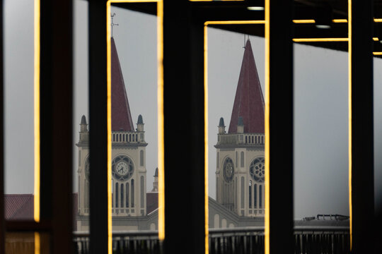 Church towers with red spires seen through dark geometric window frames illuminated by warm linear lights, creating an abstract architectural composition.