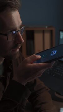 Close up of man wearing glasses talking to AI chatbot on smartphone while working at computer with artificial intelligence agent. Man using artificial intelligence assistant for work. Vertical video