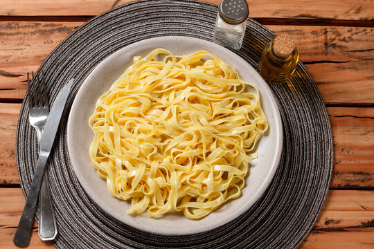 Cooked fresh fettuccine served on a rustic plate on a wooden table, accompanied by olive oil and salt, with a fork and knife alongside.
