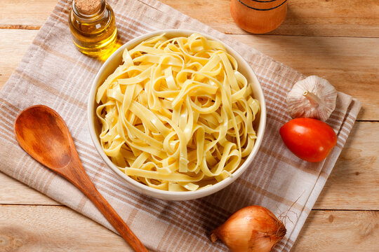 Cooked fresh fettuccine served in a bowl on a cloth napkin and wooden table, with olive oil, garlic, tomato, and onion.