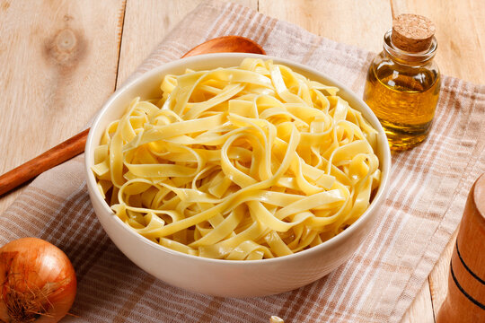 Prepared fettuccine pasta served in a bowl on a rustic cloth and wooden surface, with olive oil, garlic, tomato, and onion.
