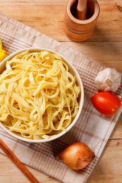 Cooked fettuccine-style pasta served in a bowl on a rustic wooden surface, with a cloth and Mediterranean culinary ingredients.