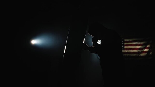 Boxer training in dark gym with dramatic lighting and American flag in background, dynamic footage of powerful punches on punching bag.