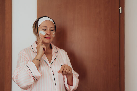 Woman in soft pink pajamas applies face cream during gentle home skincare routine creating warm lifestyle concept for mature skin moisturizing anti aging self care and beauty ritual