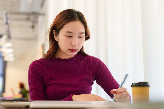 Young Asian businesswoman writing notes with coffee at desk in modern corporate office setting