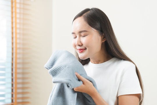Feel softness, hygiene. Smile asian young woman touch fluffy white shirt smelling fresh clean pile, stack clothes, comfort sniff after washing laundry. Household work at home, chore of maid concept.