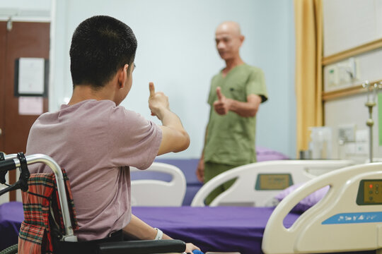 Young man in a wheelchair giving a thumbs up to a healthcare worker in a hospital room, showing support, encouragement, communication and optimism during treatment and recovery.