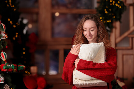 cute young woman with curly hair wears a warm red sweater while embracing a present near a glowing tree during a peaceful festive celebration