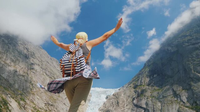Woman hiker with backpack standing with open arms against background of high mountains and glacier in Norway.