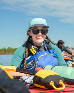 Smiling young woman with braids, cap and sunglasses sitting in a sea kayak on a paddling trip, blue sky copy space, Georgian Bay, Ontario