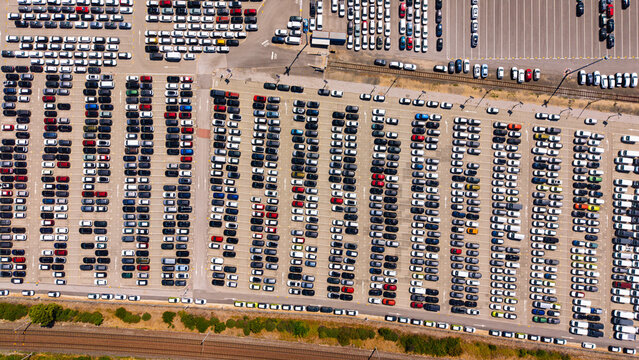 Rows of parked cars beside railway tracks, overhead view. Vehicles form a rectangular pattern on gray pavement, with red and blue cars among them.