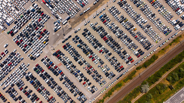 Car storage yard and railway corridor, diagonal aerial view. Dense vehicle rows border a strip of grass, with rail tracks running along the compound.