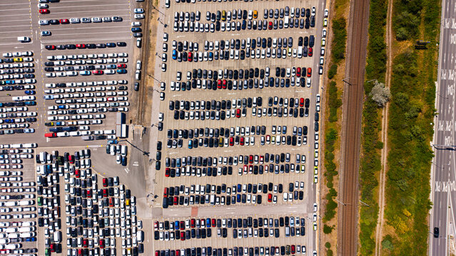 High angle view of organized car rows in an outdoor distribution hub. Hundreds of passenger vehicles sit parked on a paved lot adjacent to railroad tracks.