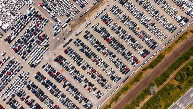Vehicle storage rows along railway tracks, diagonal aerial view. Cars form repeated bands across the pavement beside a grassy strip and a road.