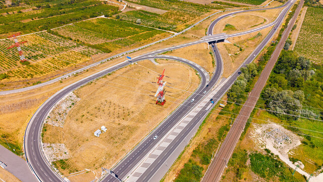 Road interchange around electricity pylon, aerial view. Looping ramps surround dry grass beside parallel rail tracks and overhead cables.