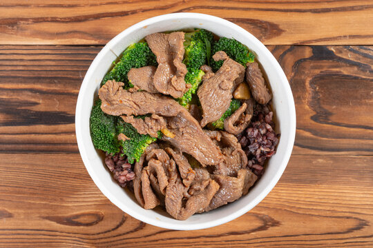 Beef grain rice bowl with broccoli in white disposable bowl on wooden table