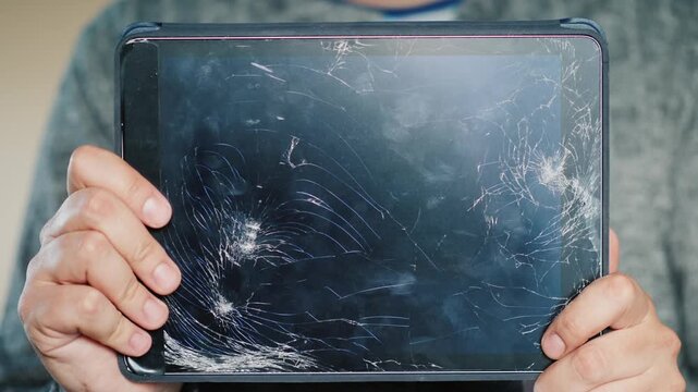 A person holds up a broken tablet with a shattered screen, showcasing the damage in a close-up shot.
