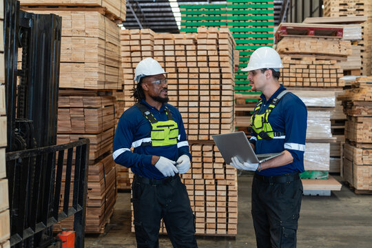 Two warehouse colleagues check lumber inventory while discussing an open laptop beside stacked wood and a forklift mast.