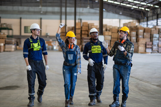 Inside the factory, four workers discuss teamwork during a walking team meeting with a tablet beside boxes and pallets.
