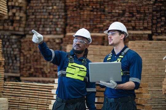 Lumber stacks surround warehouse colleagues checking inventory as one points into the distance while the other holds a laptop.