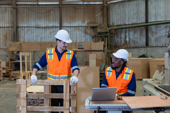 In factory teamwork, two workers hold a team meeting beside an open laptop and stacked wood in the lumber warehouse.