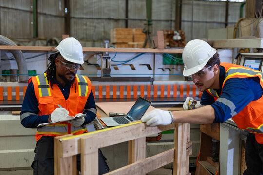 Wood workshop: woodworking carpenter checks plywood dimensions with a tape measure while his colleague writes on a clipboard.