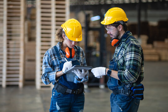 Checking workshop carpentry details, a woman reviews a tablet with her male colleague beside stacks of wooden pallets.