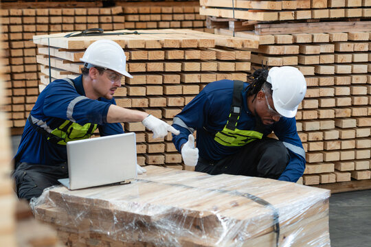 Two warehouse colleagues check lumber inventory as a man points and his coworker gives a thumbs up beside a laptop.