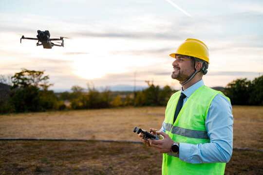 Man operates a drone while wearing a hard hat and safety vest at sunset
