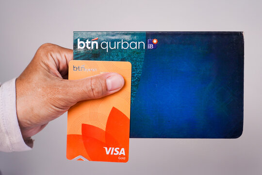 Makassar, Indonesia, September 20, A person's hand holding a BTN Qurban iB sharia savings book along with an orange BTN Visa Gold debit card against a plain light gray background.