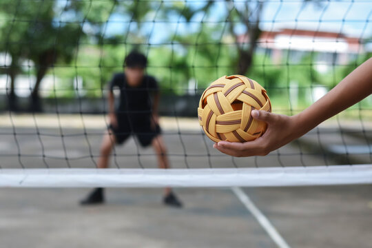 A hand holds a rattan ball at the net, ready to serve in a competitive sepak takraw game.