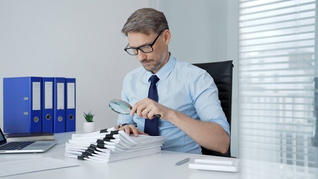 Mature businessman investigating financial documents with a magnifying glass. Performing detailed analysis. Scrutinizing reports