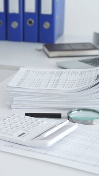 White office desk with calculator, magnifying glass, laptop showing spreadsheets and a tall stack of financial reports and binders for budgeting, tax prep and business analysis