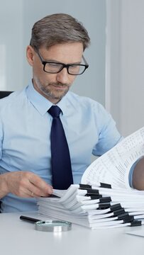 Businessman reviewing and organizing a large stack of financial reports, invoices and ledgers on a white desk with laptop spreadsheets, calculator and magnifying glass close up