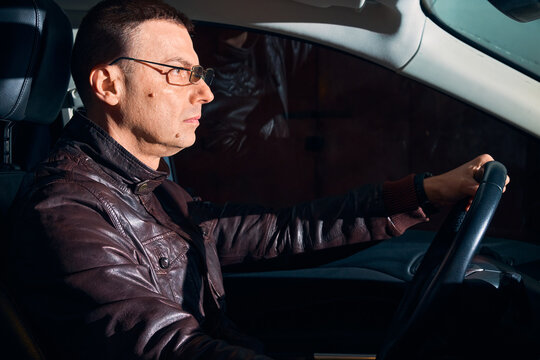 Serious mature man in glasses driving a car at night, side view through the window