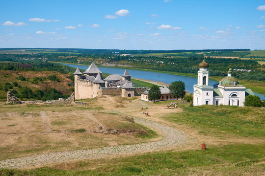 Historic medieval fortress and church overlooking a winding river and rural landscape under a bright blue sky.