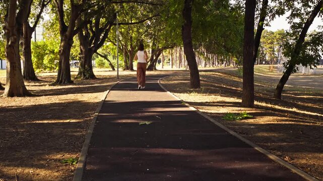 A mature female runs along a paved path under green trees in a sunny park during an outdoor workout, wide rear view.
