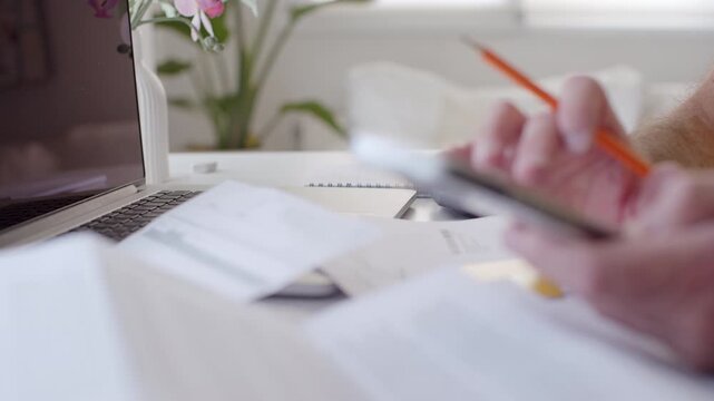 Busy person using a calculator to manage finances, checking receipts and bills while working on a laptop computer at a desk, representing the concept of budgeting and managing expenses
