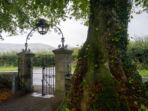 27th September 2026, Embleton, Cumbria. An ancient, moss-covered tree stands guard beside a grand stone gatehouse with an ornate lantern, hinting at the historic Saint Cuthberts Church beyond.